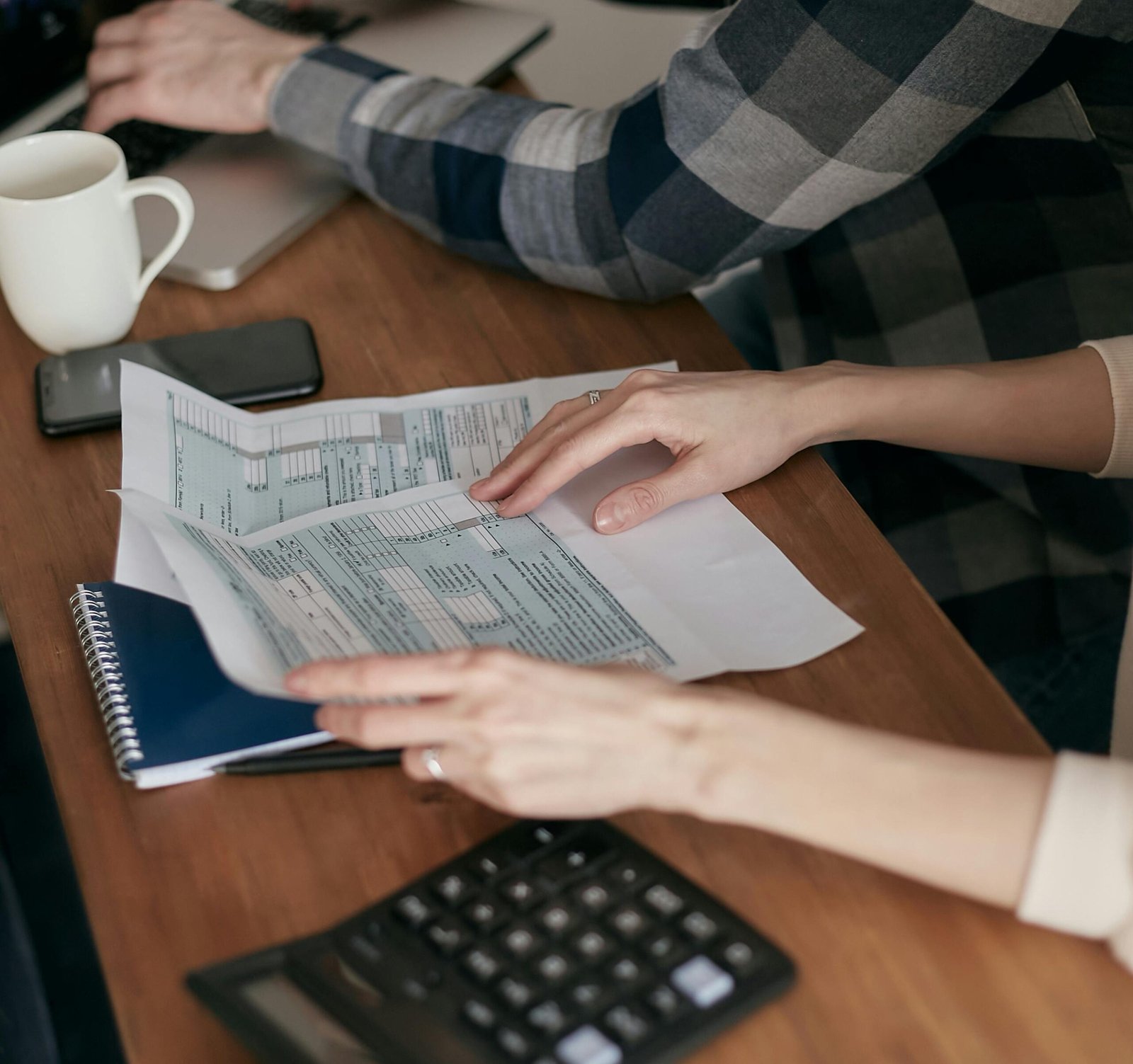 Close-up of hands organizing tax forms on a desk with a calculator, laptop, and notebook.